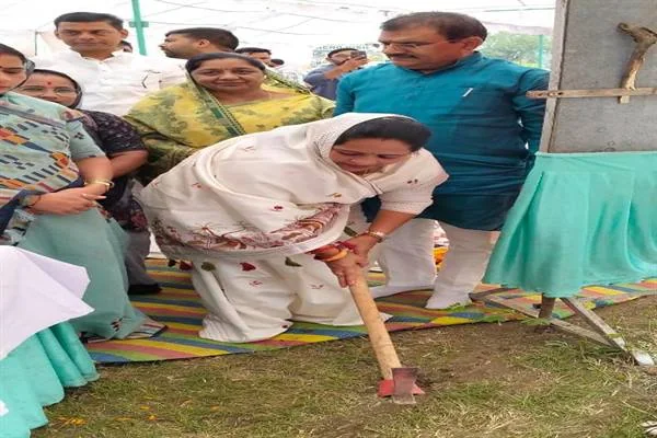 सफेद साड़ी पहनी महिला भूमि पूजन स्थल पर कुदाल चलाती हुईं, आसपास खड़े जनप्रतिनिधि और अधिकारी